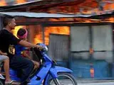 Una familia timorense sobre una motocicleta pasa por delante de una casa ardiendo en Dili, Timor Oriental. (Foto: Efe)