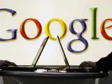 Dos personas usan sus portátiles con el logo de Google de fondo.