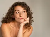Portrait of thoughtful young woman thinking with hand on cheek against grey background. Beautiful natural girl with bare shoulder looking away with funny expression. Close up face of pensive beauty woman with copy space.