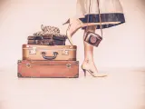 Woman standing with her vintage suitcases inside a home concept symbolizing she is going away on a trip or going on a vacation. Woman is wearing a beige dress and shoes. She is holding a vintage camera. Only showing the woman's legs, hands and shoes. Horizontal sepia image. Copy space.
