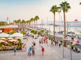 People walk on the restaurant and palm tree lined Paseo del Mullene Uno waterfront promenade in downtown Malaga Spain, at sunset time, with a docked cruise ship in the background